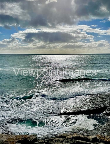 Sparkling blue ocean waves crashing against a rocky shore under a cloudy sky with bright sunlight.