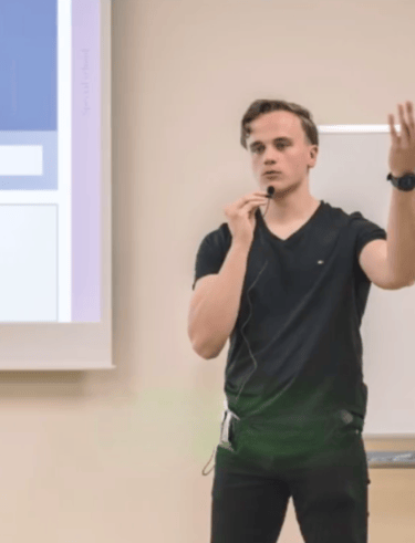 Julius-Sensei steht in einem Klassenraum vor einer Projektionsfläche, hält ein Mikrofon und erklärt gestikulierend ein Thema