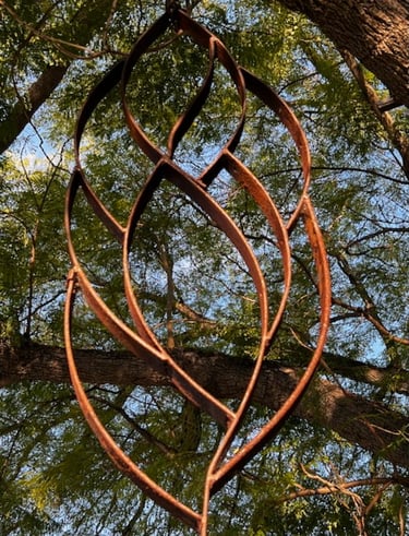 Rusty iron metal garden sculpture hanging from a large tree with green leaves.