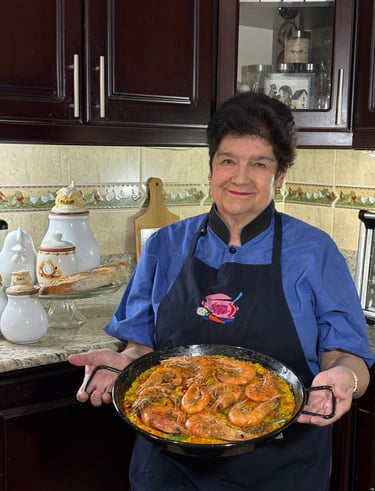 A smiling chef in a blue shirt holding a fresh seafood paella with large shrimp in a traditional pan.