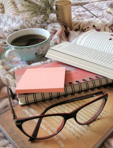 A pair of glases, a novel, a cup of tea, and a notebook sit on a shabby-chic tablecloth.