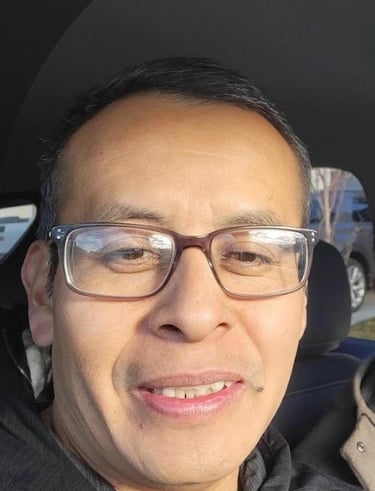 A smiling man wearing rectangular eyeglasses and a black hoodie sitting in a car.