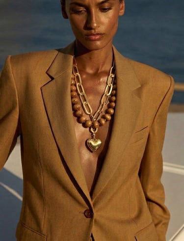 A woman wears a tan linen blazer and gold chain necklaces with a heart pendant by the ocean.