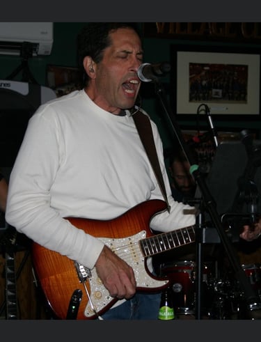 A male musician singing into a microphone while playing a sunburst electric guitar during a live band performance.
