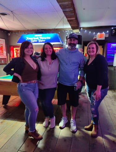 Group of friends smiling in a sports bar next to a pool table with neon beer signs.
