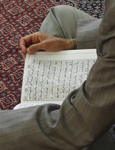 A person sitting on a patterned rug while reading the Holy Quran with Arabic calligraphy.