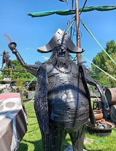 A large black metal pirate sculpture made of scrap metal standing outdoors in front of a ship mast.