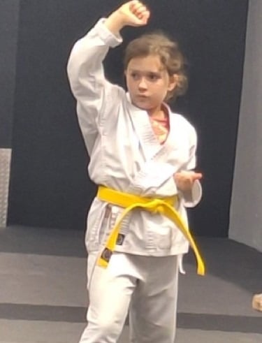 Young girl in a white karate gi with a yellow belt practicing a high block technique in a martial arts dojo.