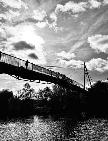 Black and white silhouette of a suspension bridge over a river with a cloudy sky backdrop.