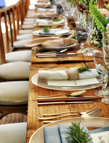 Rustic wedding table setting with gold cutlery, glass chargers, and rosemary sprigs on a wood farm table.