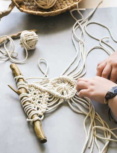 Manos elaborando un colgante de macramé DIY usando cuerda de algodón blanca y un palo de madera