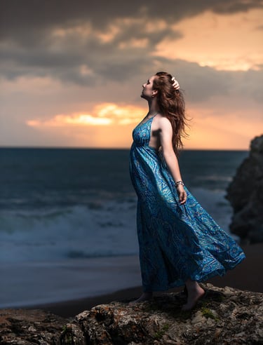 Creative portrait of a woman in a blue dress standing on rocks at sunset