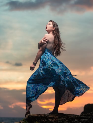 Portrait créatif d’une femme en robe bleue posant sur des rochers au coucher du soleil.