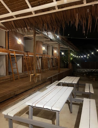 Rustic outdoor hostel lounge at night with white picnic tables and wooden sleeping pods.