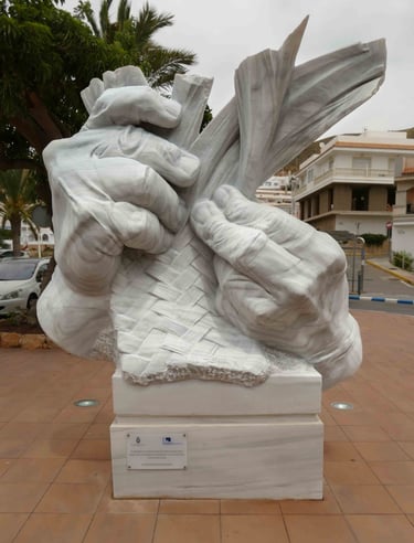 White marble sculpture of hands weaving a rope braid on a pedestal in a city square.