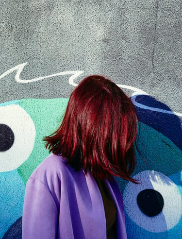 A woman with vibrant red hair wearing a purple coat stands against a blue graffiti wall.