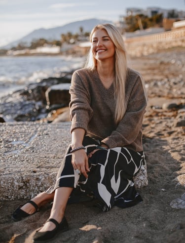 Smiling blonde woman in a brown v-neck sweater and patterned skirt sitting on a beach at sunset. 