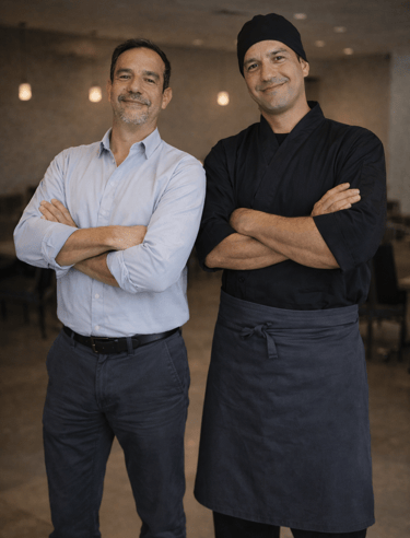 Chef Jairo e Fernando, sócios do Okone Campo Belo, posando sorrindo com braços cruzados no salão do restaurante.