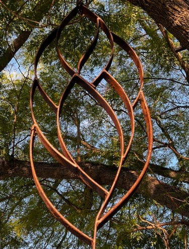 Rusty iron metal garden sculpture hanging from a large tree with green leaves.