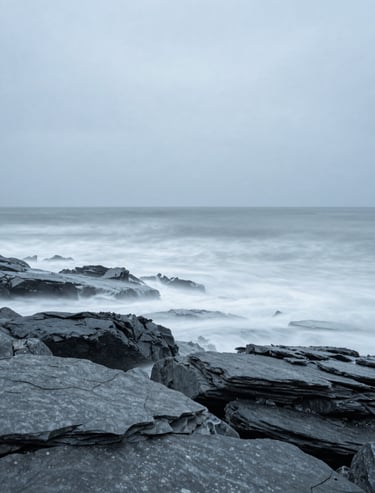 A minimalist long exposure shot of waves hitting a slate gray rocky shore. Soft misty texture, light blue gray sky, Global / General coastal scene.