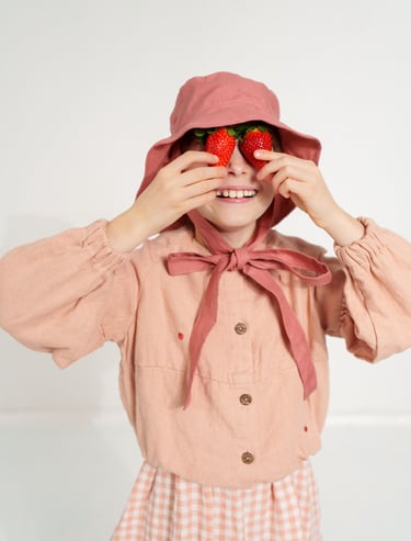 Smiling child in a dusty pink bonnet and dress playfully holding two strawberries over their eyes, d