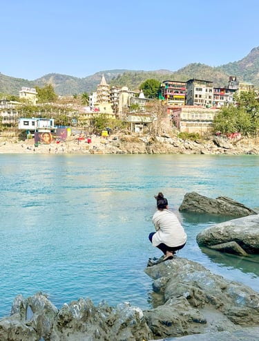 Eva standing by the sacred Ganges River in Rishikesh, India, with mountains visible in the background on a sunny day.