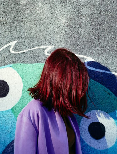 A woman with vibrant red hair wearing a purple coat stands against a blue graffiti wall.