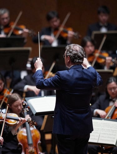A dramatic shot of the orchestra pit from above, with the conductor's baton raised. Colors include deep #2B2D42 and orchestral highlights in #C8B8B8.