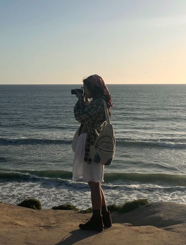 Vibey full body candid shot of me holding camera up to my face, standing on cliffs over ocean.Earthy