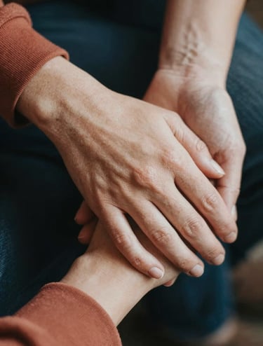 A detailed close-up shot of two people's hands gently intertwined. The lighting is soft and natural, highlighting skin textures and a terracotta-toned sleeve. The mood is intimate and storytelling-focused.