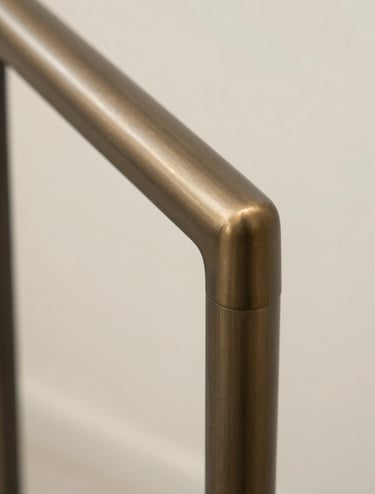 Macro shot of a staircase handrail detail made of muted bronze. The texture is smooth with a satin finish. The background is a soft-focus soft off-white, highlighting the warm minimalism of the hardware.
