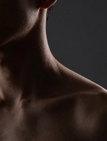 A close-up of a neck and shoulder, captured in low-light. The light catches the edge of the skin, creating a thin off-white highlight against a deep dark gray background. Sophisticated and moody North American / US photography.