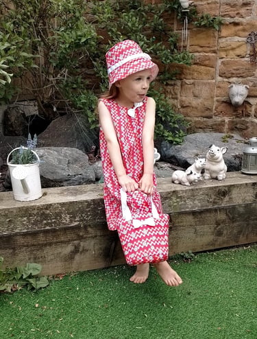 Young girl in a red and white patterned sun hat and dress holding a matching bag in a garden.