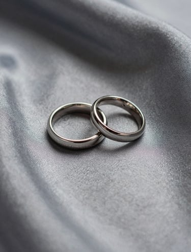 A minimalist macro photography shot of two wedding bands resting on a soft silver fabric. Professional studio lighting with deep dark charcoal shadows. South American / Colombian wedding context.
