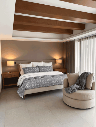 Recámara con acabados cálidos, tapiz neutro, cama con textiles grises y sillón individual | KARÄNA