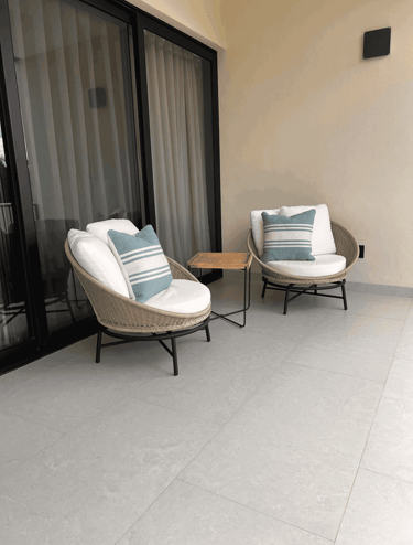 Sillas de mimbre con cojines blancos y mesa lateral en terraza con ventanal de cristal | KARÄNA