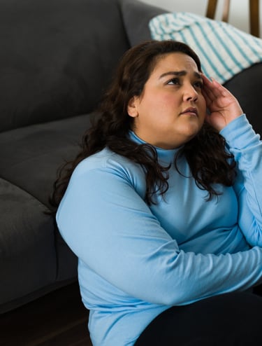 A stressed woman sitting on the floor leaning against a gray sofa, suffering from a painful headache.