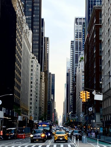 A bustling New York City streetscape with tall skyscrapers, traffic, and pedestrians under a bright sky.