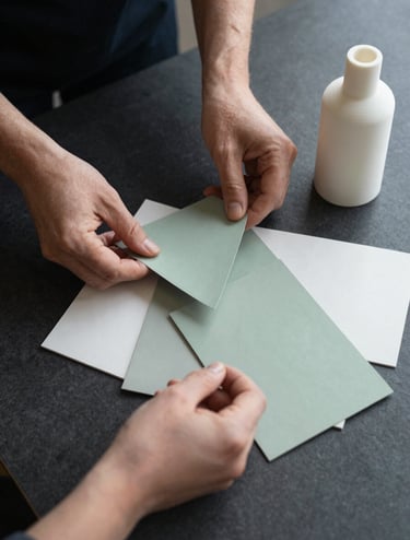 A top-down view of a professional's hands in a North American / US studio, organizing various paper swatches in soft sage green and mist white alongside a 3D-printed bottle prototype on a dark charcoal desk.