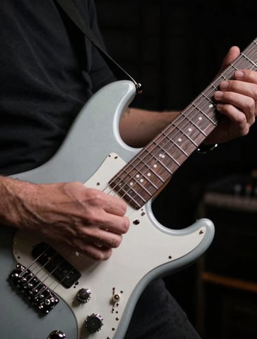 Close-up detail of a professional musician's hands on a sleek electric guitar in a dark, atmospheric studio. The lighting is focused and artistic, using light gray highlights on metallic parts against a black background. North American / US vibe.
