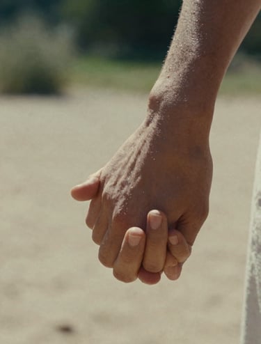 Close-up photography of two hands intertwined, sunlight catching the skin. Cinematic film style with a shallow depth of field. Soft sand and warm skin tones, set in a European / French garden. Storytelling focus on intimacy.