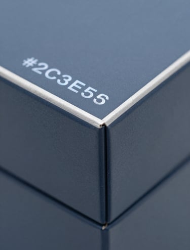 A macro photograph showing the corner of a luxury perfume box. The packaging features a matte finish in #2C3E50 with a silver-blue #8D9FB5 foil stamp. Clean geometric lines and meticulous craftsmanship visible in the sharp paper folds. High-end, forward-thinking aesthetic.