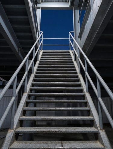 A vertical shot looking up through an industrial stairwell. The steps create a repetitive pattern of dusty grey-blue metal against a midnight blue sky seen through a skylight. The composition is highly symmetrical and professional.