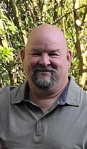 Smiling Donnie Hough with a grey goatee wearing a polo shirt outdoors against green foliage.