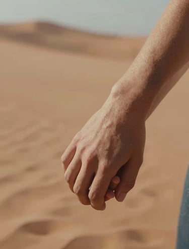 A close-up photograph of a couple's intertwined hands, with a soft-focus background of the North American / US desert. The lighting is warm and sun-drenched, featuring soft sand and muted terracotta tones to evoke a sense of deep human connection and cinematic intimacy.
