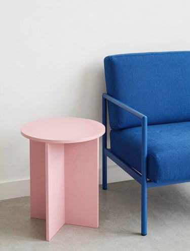 A high-angle professional photograph of minimalist Bauhaus-style furniture in a bright, modern North American apartment. The scene features a cerulean blue armchair and a soft pink side table against a clean white wall, showcasing sharp geometric lines and elegant composition.