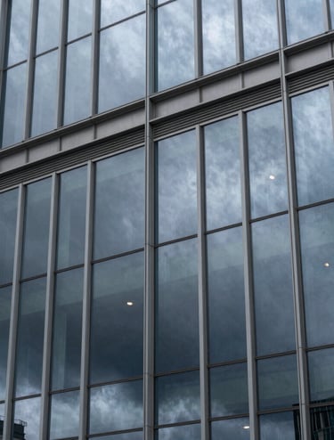 Close-up architectural photography of a modern, glass-facade building reflecting a storm sky. The palette is dominated by slate grey and cool blue tones. High-quality professional shot with clean geometric lines. International / Western city.