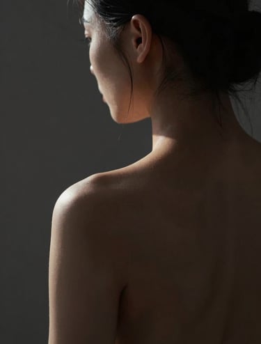 A fine art portrait in extreme chiaroscuro. The subject's back is visible, with light grazing the spine and shoulder blade, creating a sculptured effect. Background is deep charcoal #1F1E1C. The light has a warm, intimate #F5F2EF glow. Sophisticated and emotive composition.