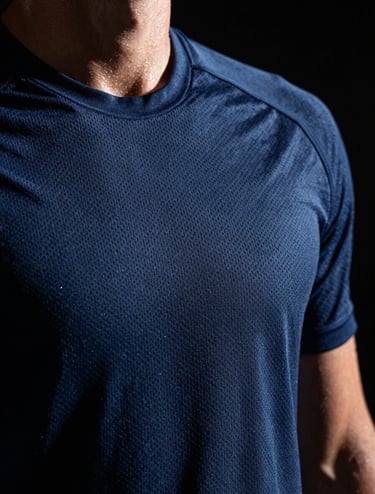 A close-up photograph of an athlete's textured dark blue mesh jersey, capturing the intricate weave pattern and droplets of sweat. Dramatic side lighting creates high contrast against a black background. Professional sports photography, Western / International context.