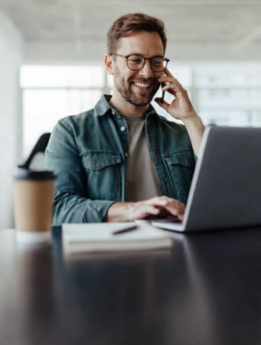 Smiling copywriter with glasses talking on smartphone while working on laptop in office.